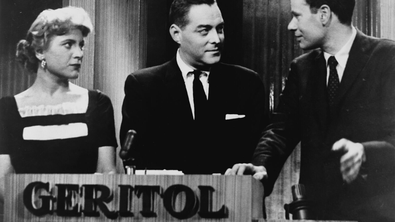 A black and white television still of three people behind a lectern marked GERITOL: a woman in a dark dress on the left, a man in a suit in the centre turning to face a younger man on the right