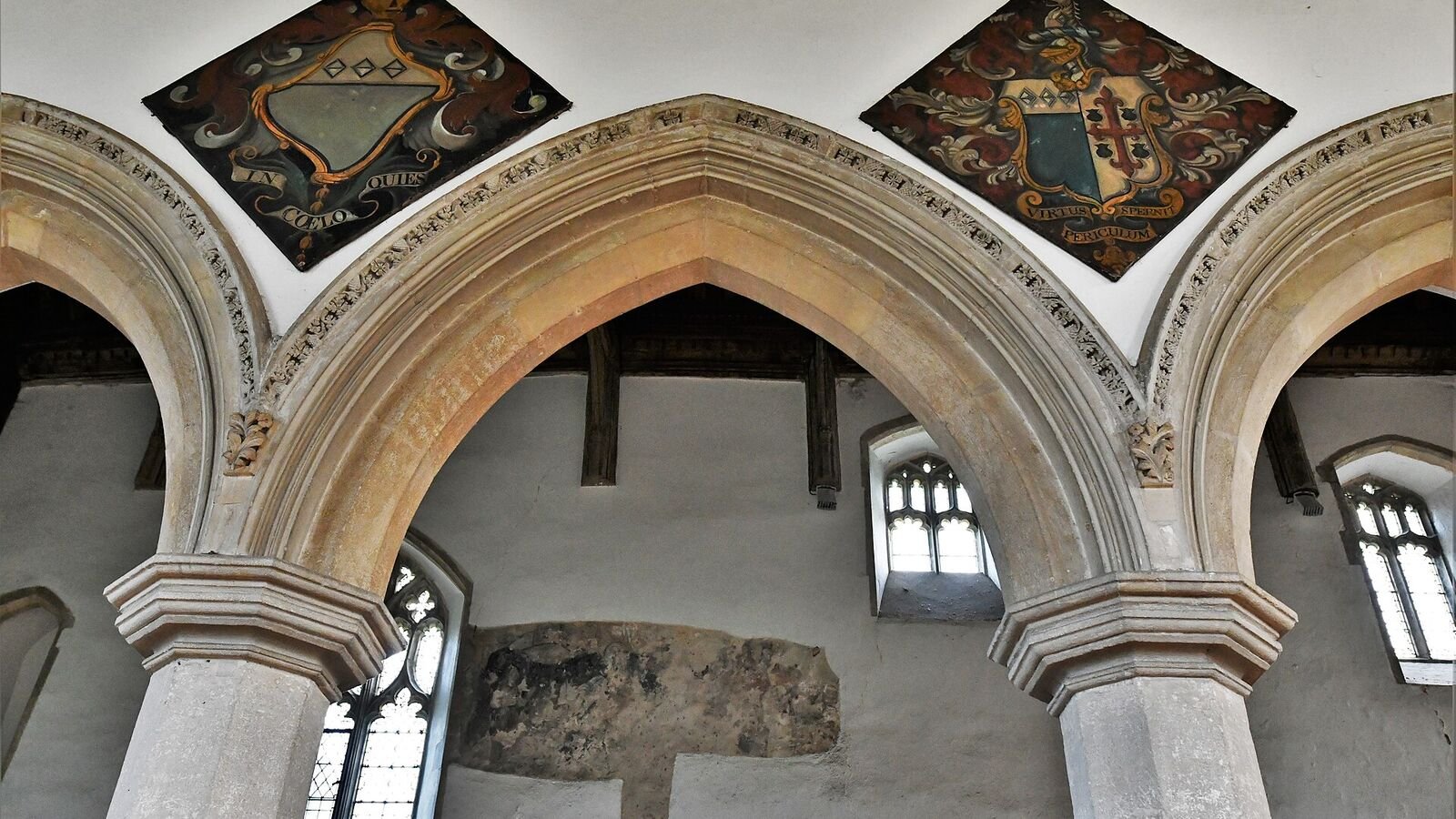 Two painted heraldic hatchments, diamond-shaped boards bearing coats of arms, mounted high on the whitewashed wall above the stone arches of a medieval church interior.