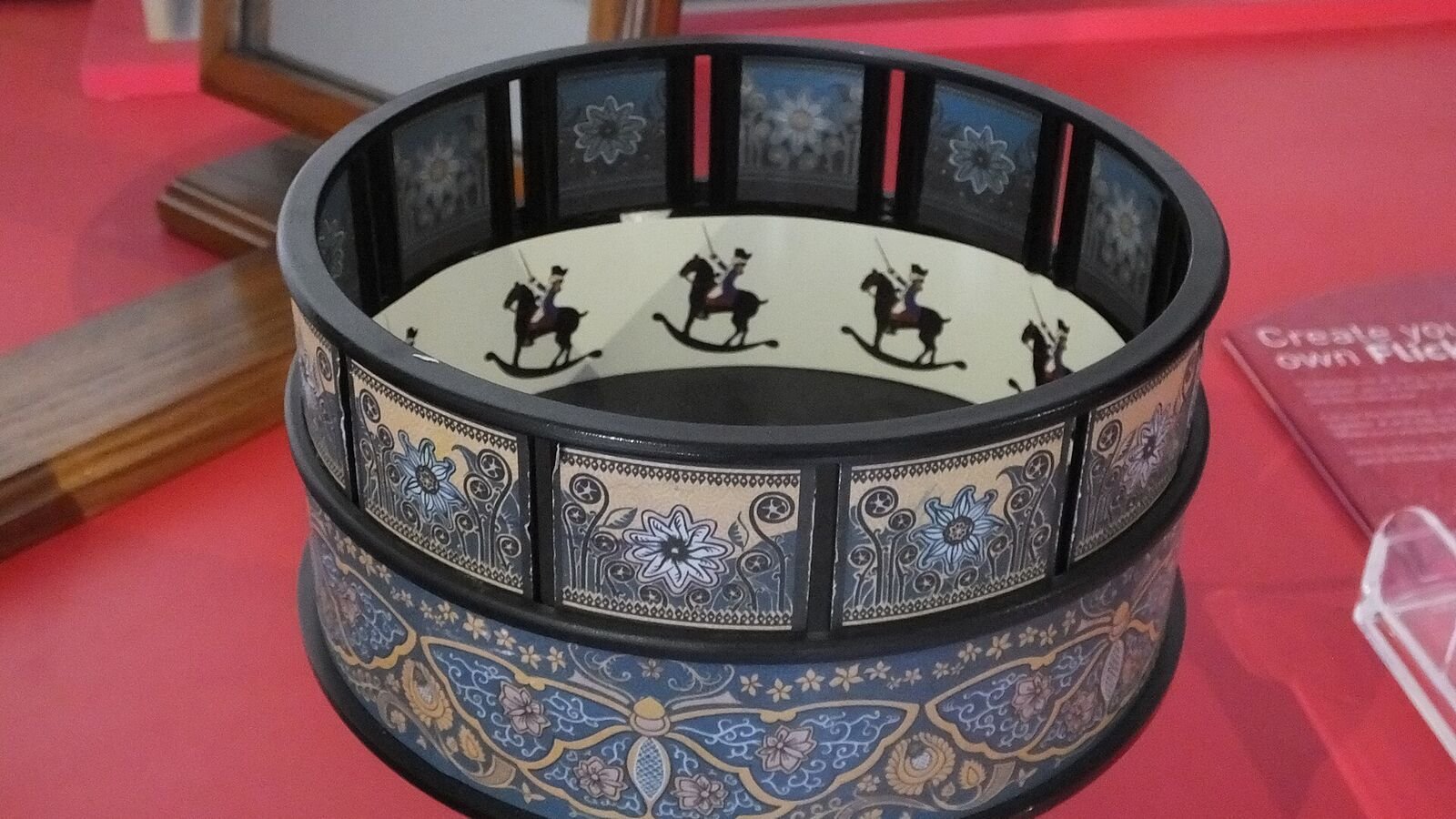 A decorated Victorian zoetrope: a black drum with vertical viewing slots and a printed floral pattern around the outside, with a paper strip of horse-and-rider drawings visible inside.
