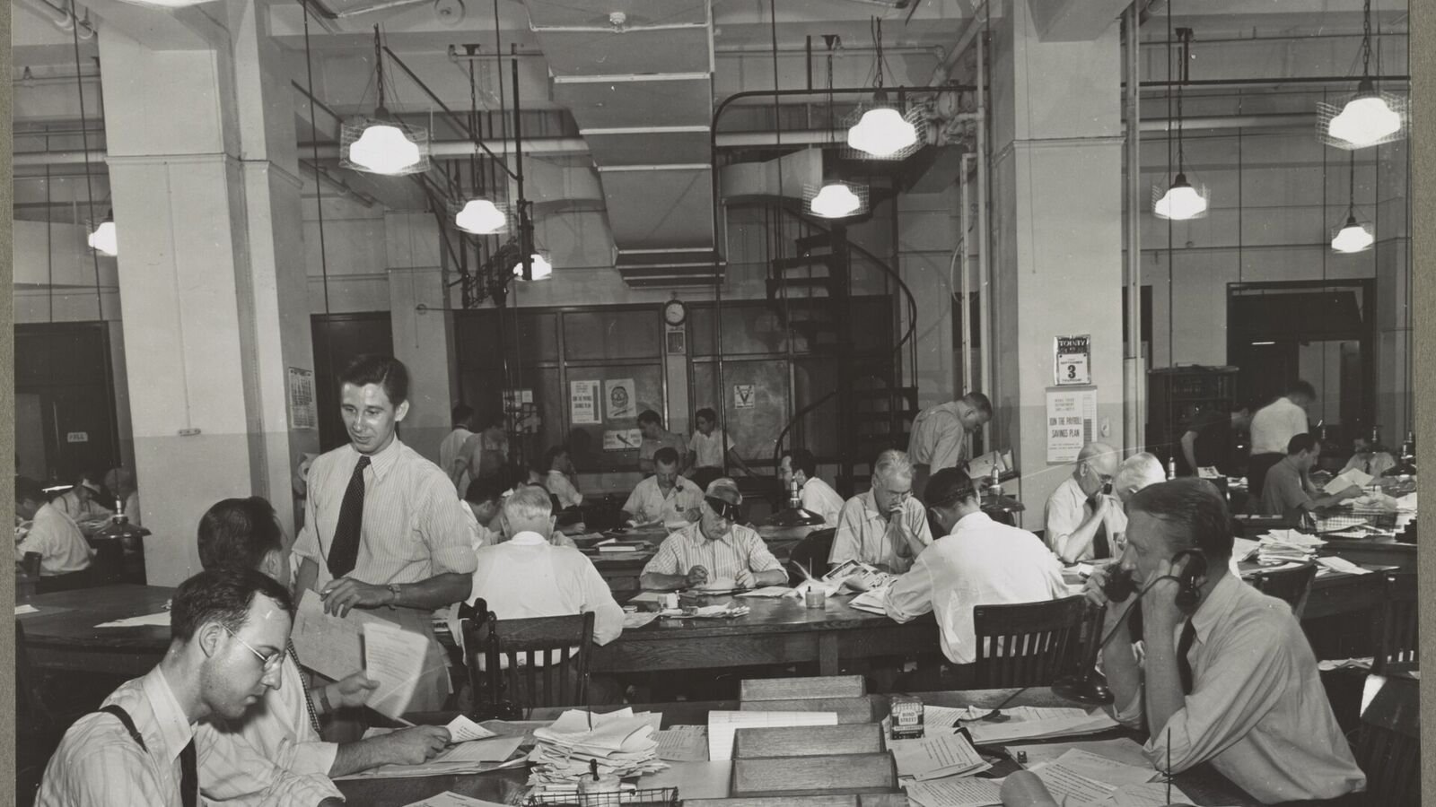 A black and white photograph of a crowded 1940s newspaper office, with rows of men in shirtsleeves at wooden desks piled with paper, some on telephones, under hanging lamps.