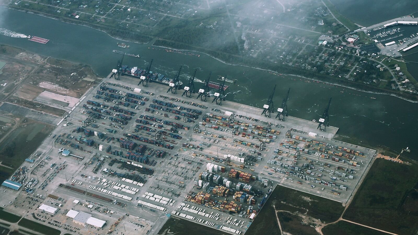 Aerial view of a container terminal beside a waterway, with rows of stacked coloured containers, lines of parked lorry trailers and a row of gantry cranes along the quay.