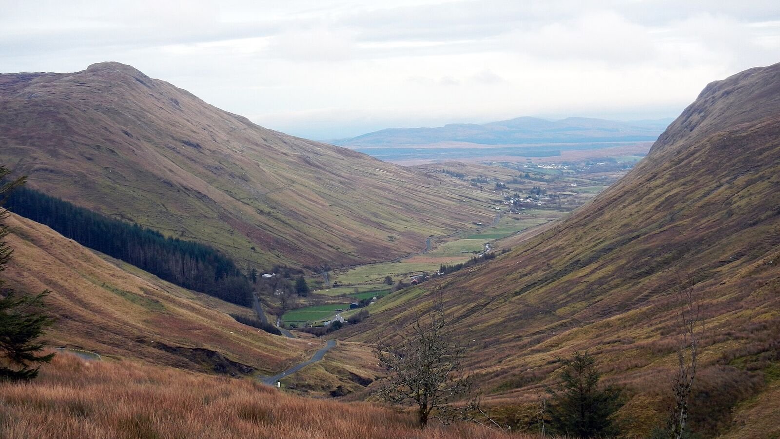 A broad glacial valley with steep grassy sides curving down to a narrow road and a small river on the flat floor, opening out towards farmland and distant hills under grey cloud.