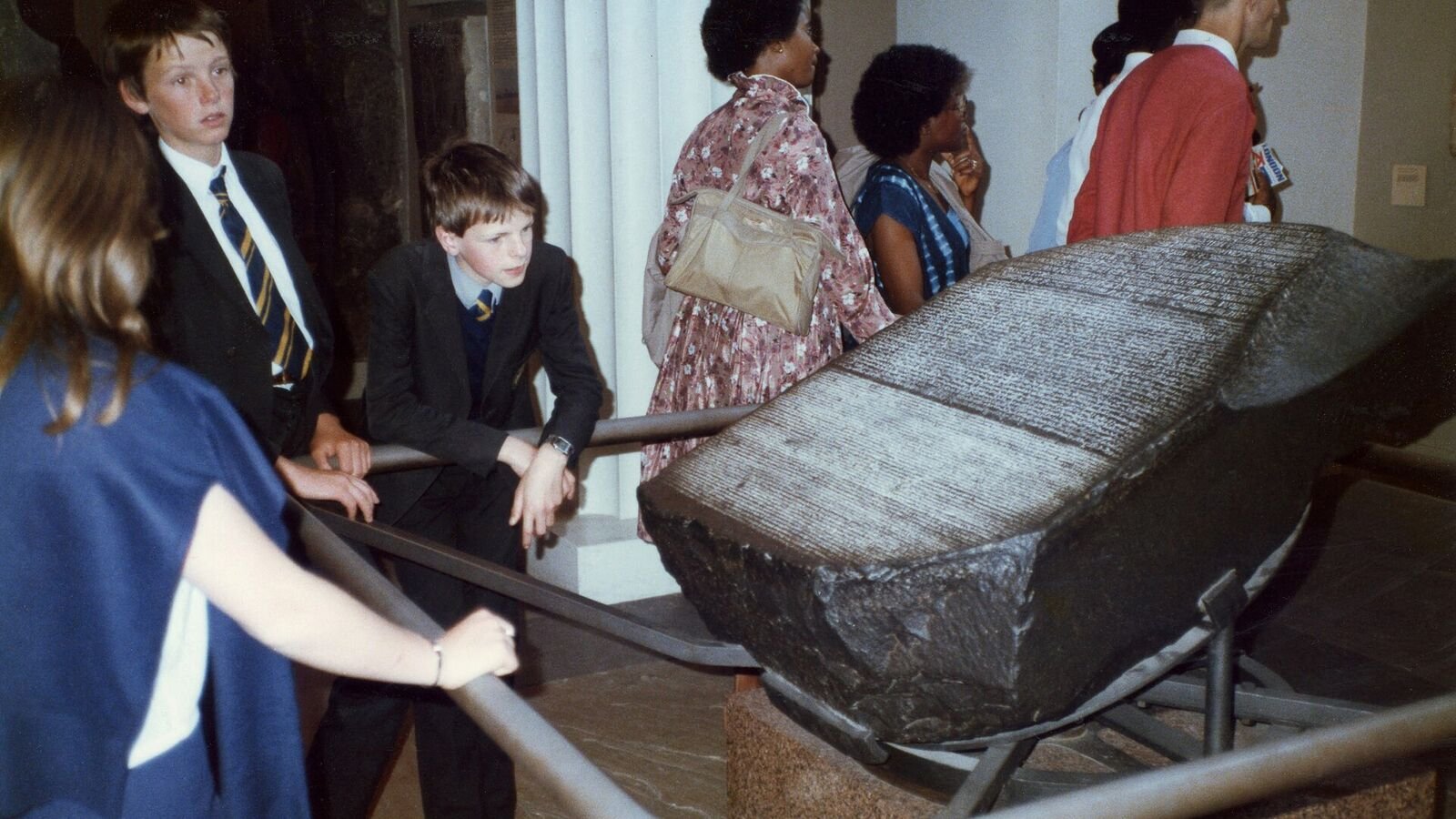 Museum visitors, including two schoolboys in blazers and ties leaning on a metal rail, gathered around the Rosetta Stone, which lies tilted on a low stand with its dense lines of carved text facing them.