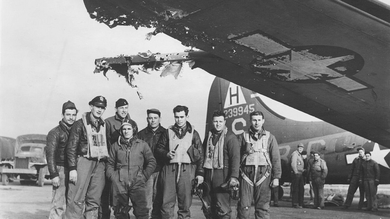 Eight American airmen in flight jackets and flying suits standing on an airfield beneath the shredded wing of a B-17 bomber, with a large section of the wing's trailing edge torn away.