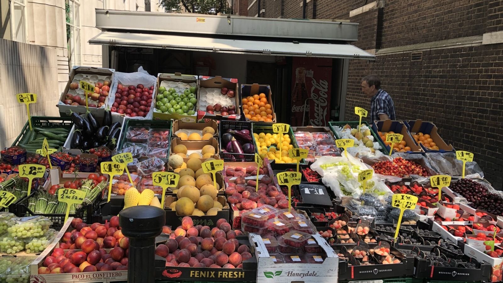 A London street fruit and vegetable stall stacked with crates of peaches, grapes, strawberries, melons and aubergines, covered in bright yellow hand-written price tags, with the stallholder standing at the far right.