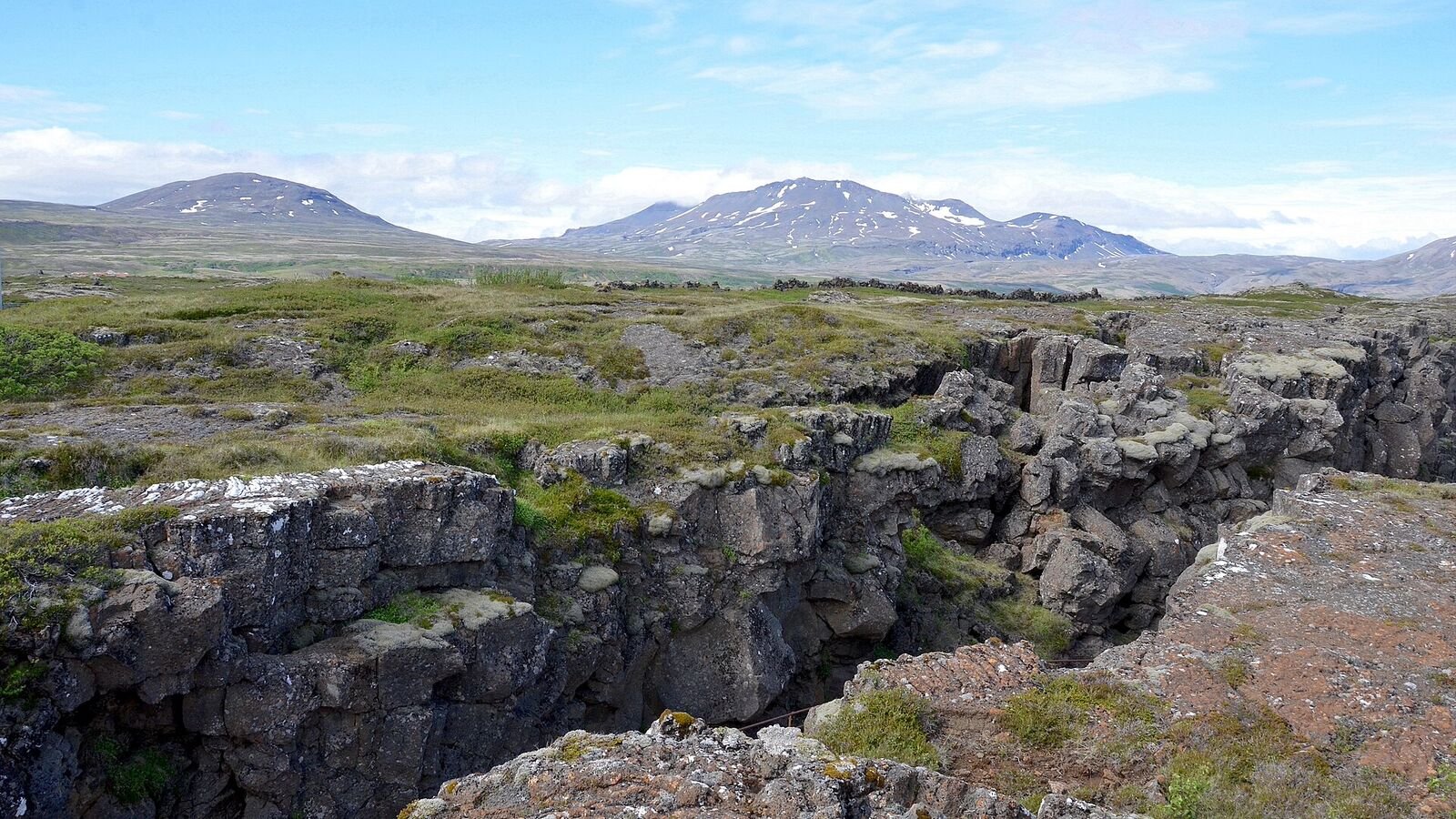 A deep, straight-sided fissure splitting a mossy volcanic plain in Iceland, with snow-streaked mountains and blue sky behind.