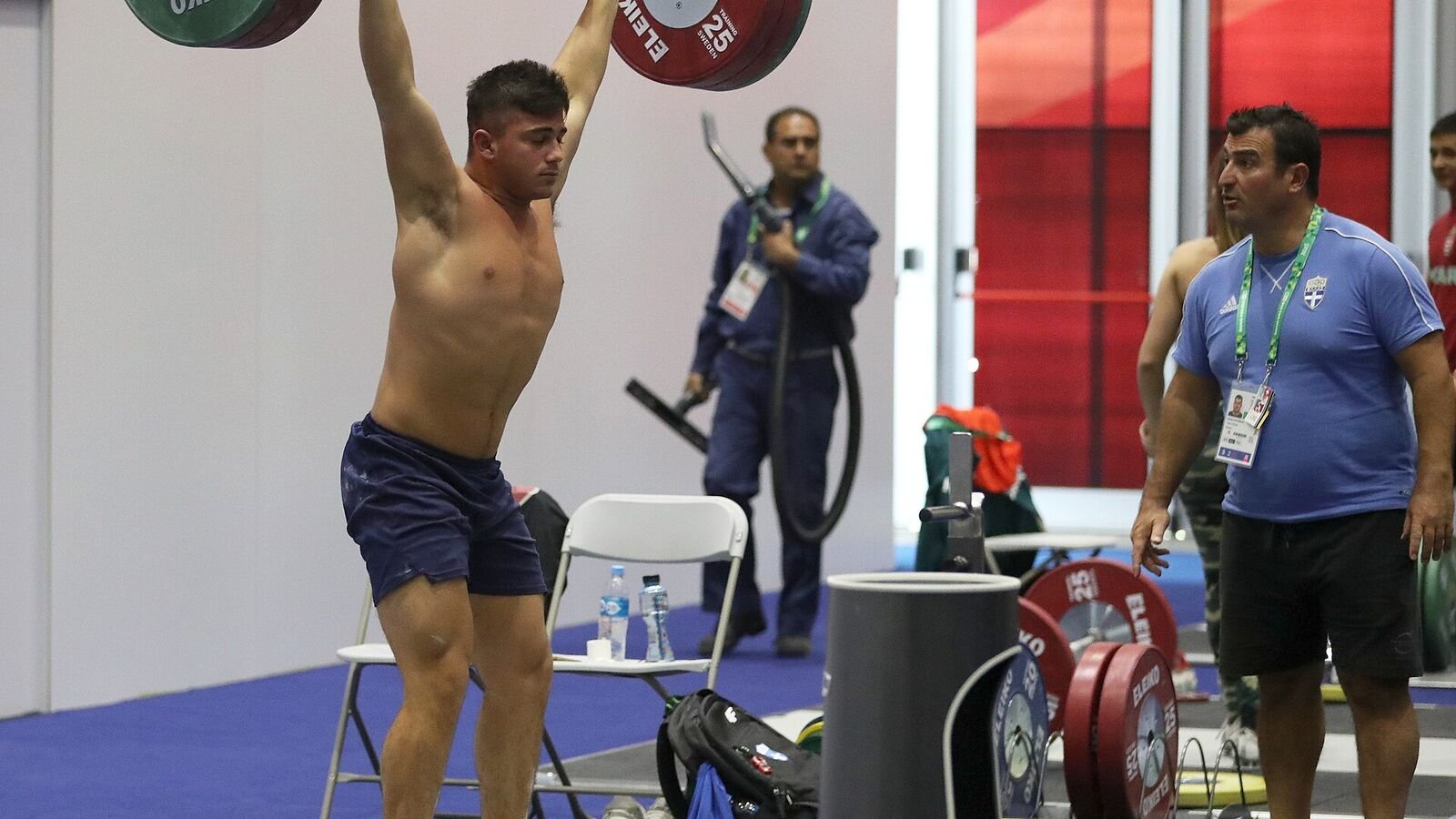 A young weightlifter in blue shorts holding a loaded barbell locked out overhead in a training hall, with coaches and stacked plates behind him.