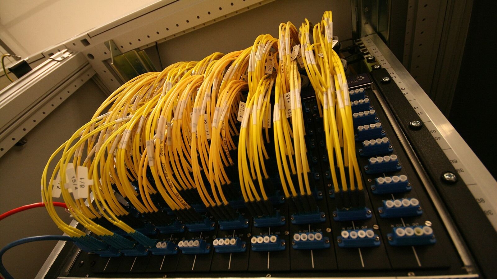 A rack-mounted optical patch panel packed with dozens of bright yellow fibre optic cables, each carrying a hand-written numbered label, curving down into rows of blue connector ports.