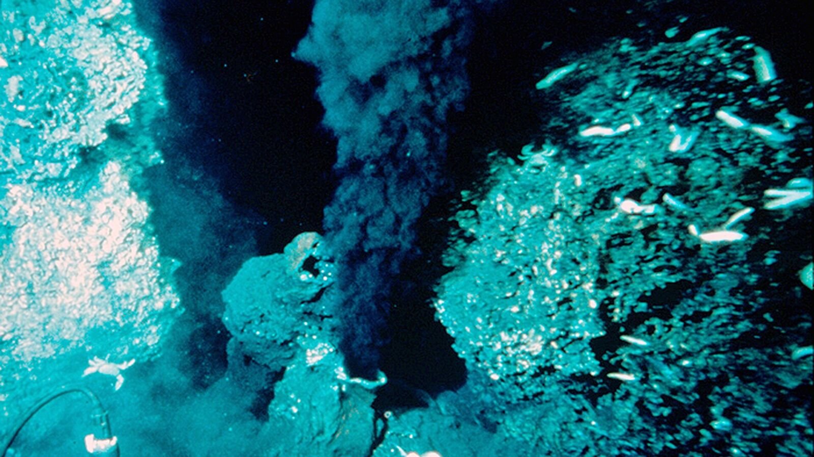 A dark hydrothermal vent chimney on the seafloor releasing a dense plume of black mineral-laden fluid into the surrounding water, lit by a submersible.