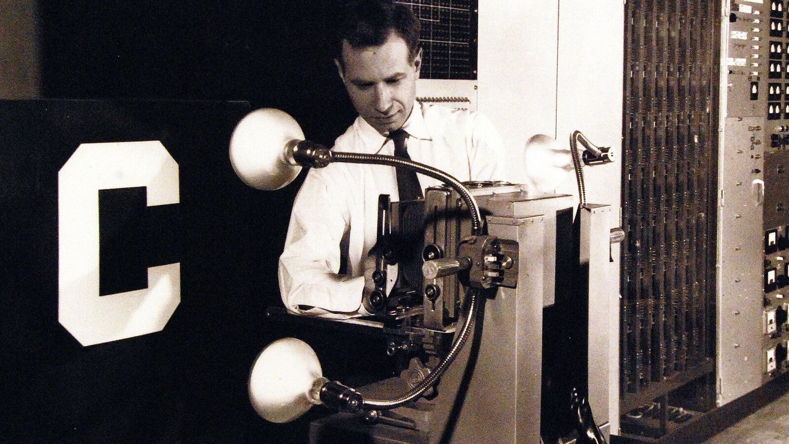 A black and white photograph from around 1960 of a man in a white shirt and dark tie leaning over the camera and lamp assembly of the Mark I Perceptron, with a large letter C panel to his left and tall racks of electrical equipment behind him.