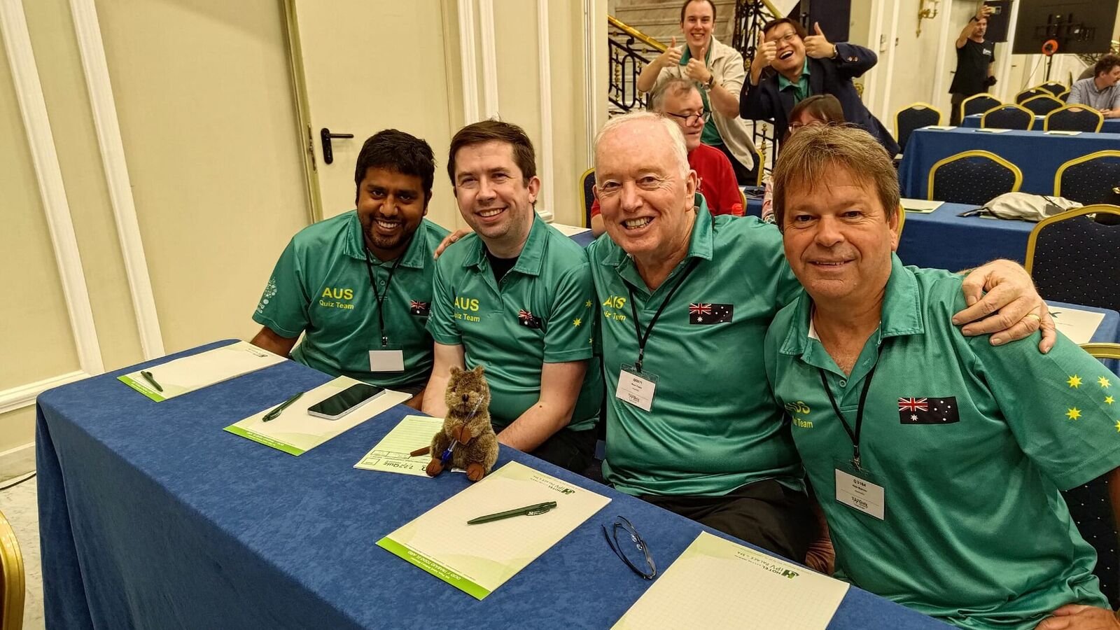 Four men in matching green Australian quiz team shirts sitting together behind a blue-clothed table with blank answer papers and pens in front of them, other competitors visible behind