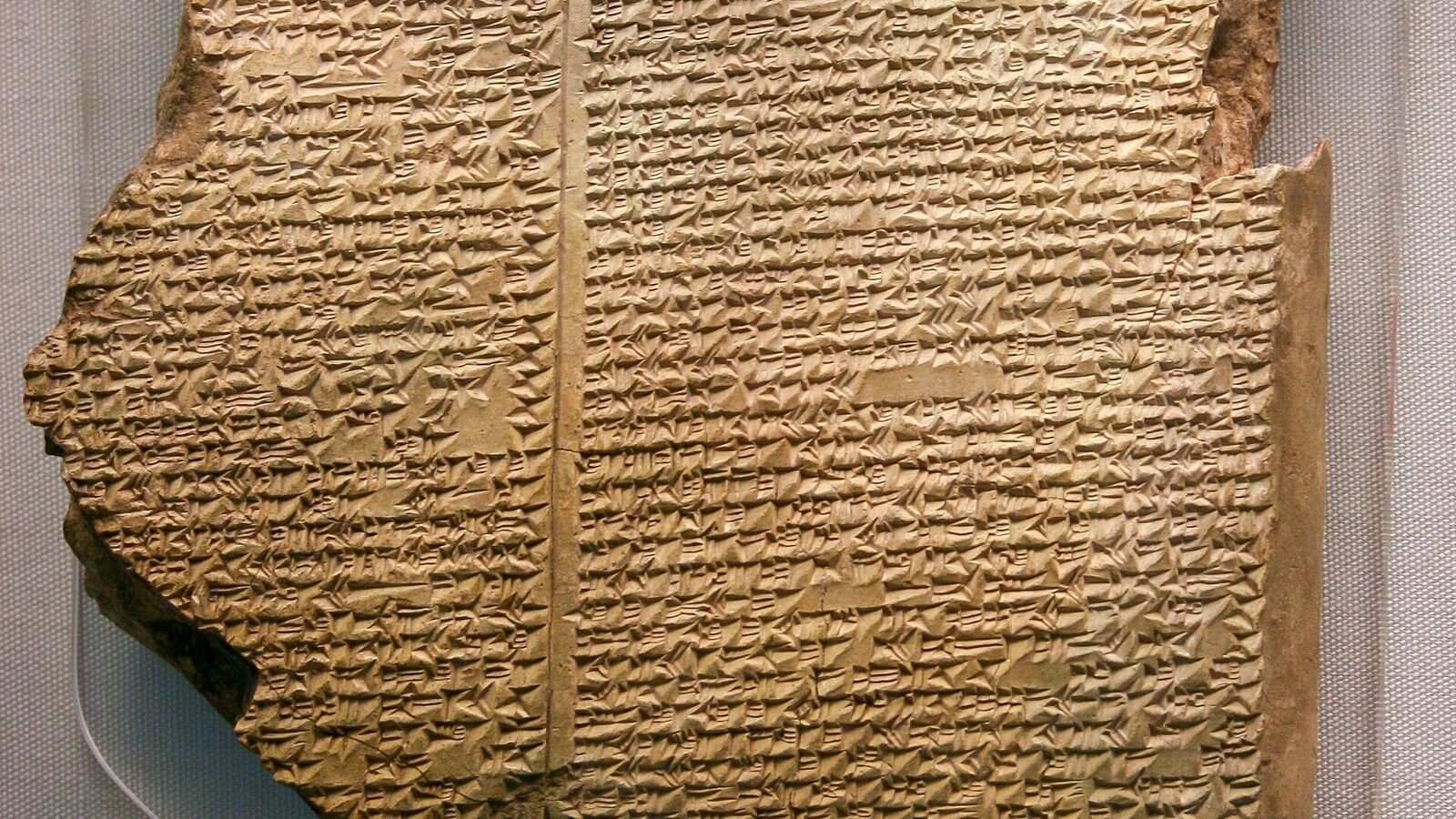 A broken clay tablet covered in dense columns of cuneiform writing, cracked along one edge, displayed in a museum case.