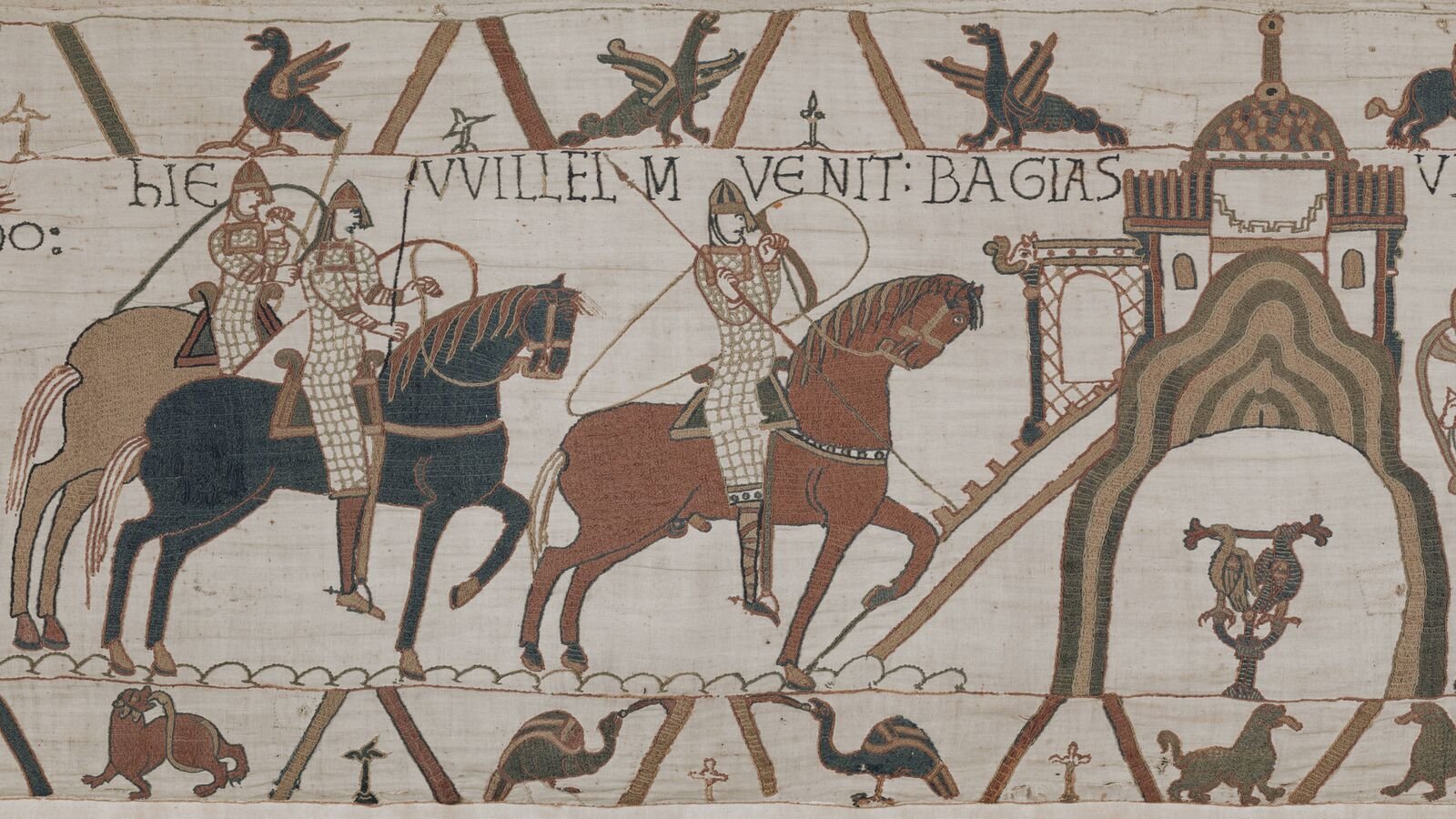 A section of the Bayeux Tapestry showing mounted Norman soldiers in mail armour riding towards a wooden tower on a raised mound, with embroidered Latin lettering above them and borders of birds and beasts.