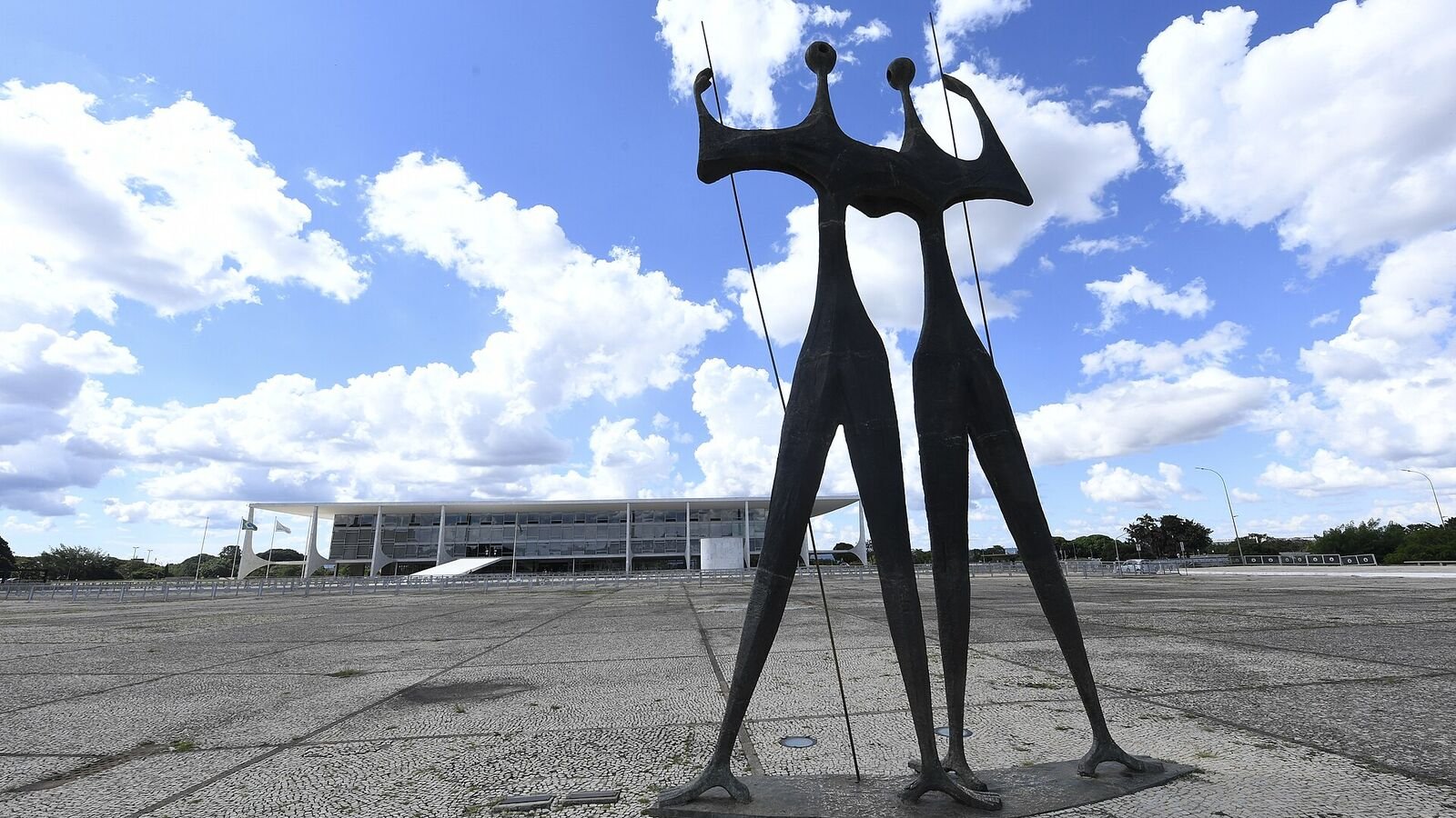 Two tall, elongated bronze figures holding poles stand on a vast paved plaza under a cloudy blue sky, with a long low white modernist palace of thin columns in the background.