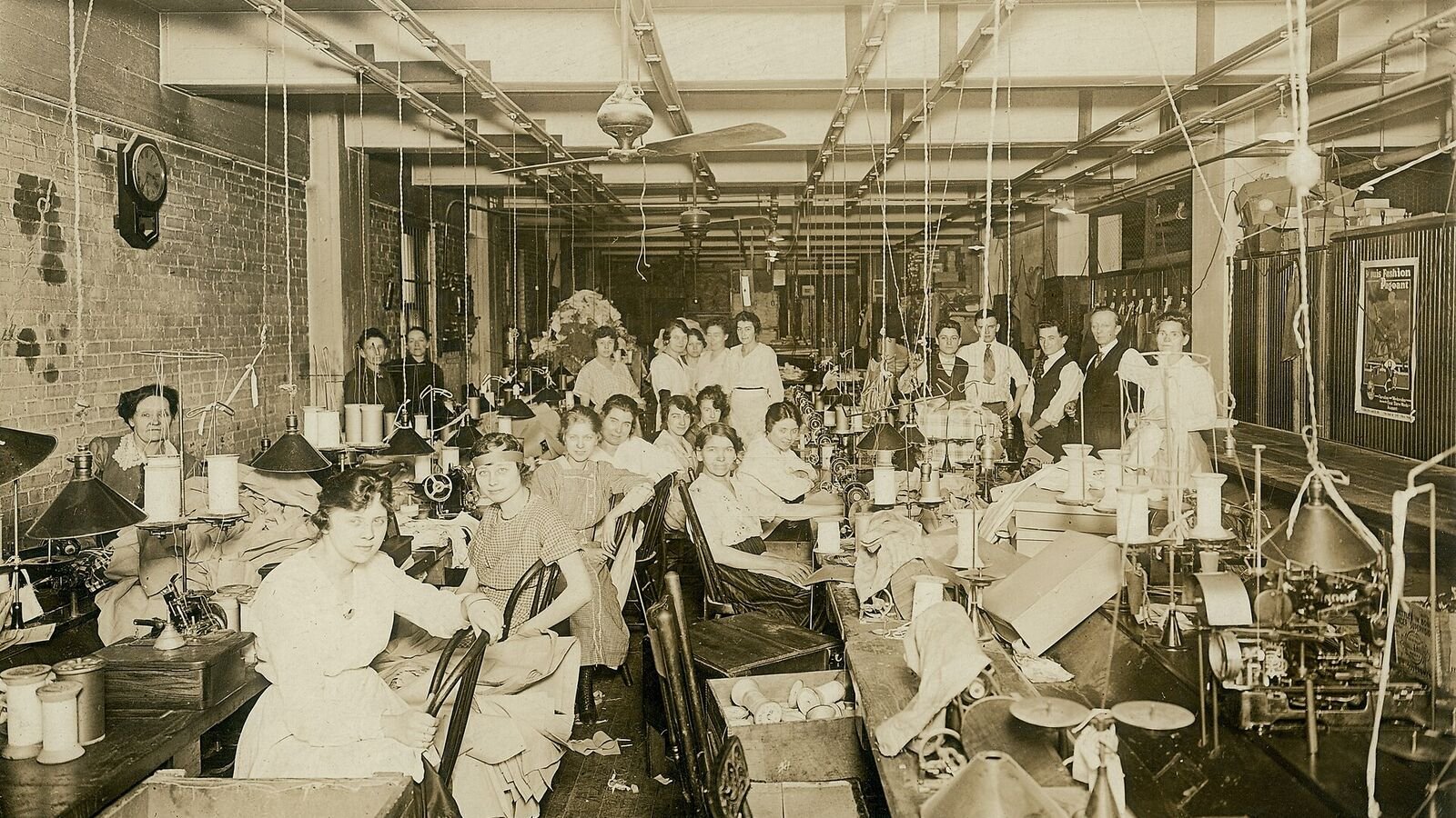 A large early twentieth-century workroom filled with rows of women seated at treadle sewing machines, with bundles of cut cloth stacked beside them.