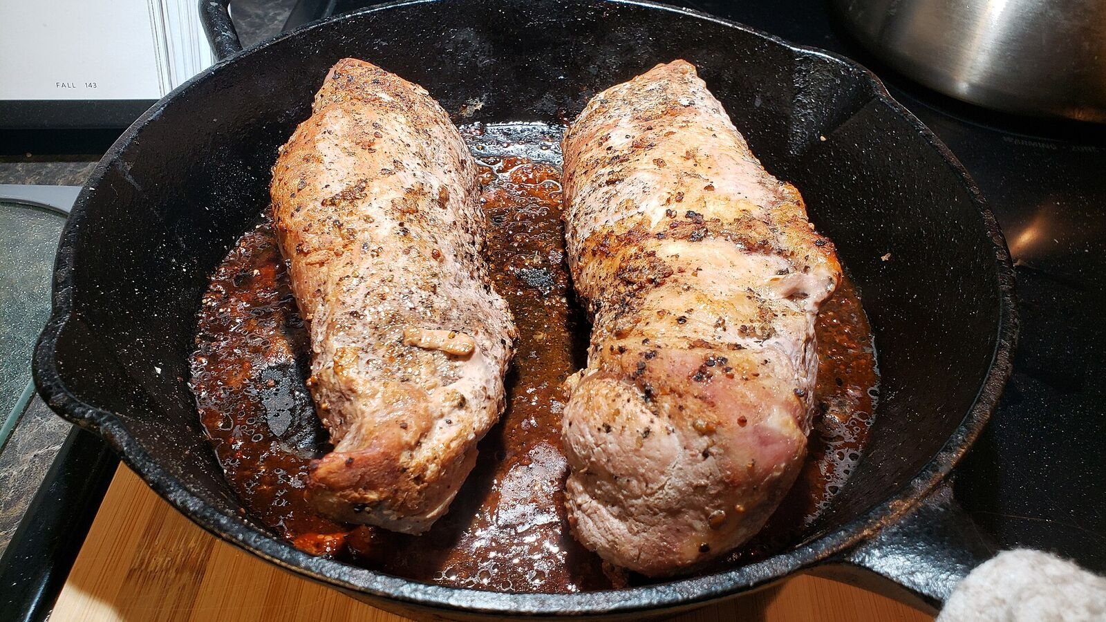 Two browned pork tenderloins resting in a black cast-iron skillet on a hob, with dark sticky juices pooled around them and a wooden board at the edge of the frame.