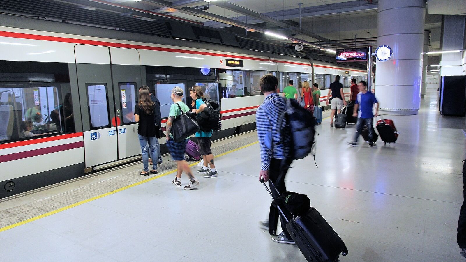 Travellers crossing a polished airport station platform towards a red and white train, several of them pulling upright wheeled suitcases behind them by extended handles.