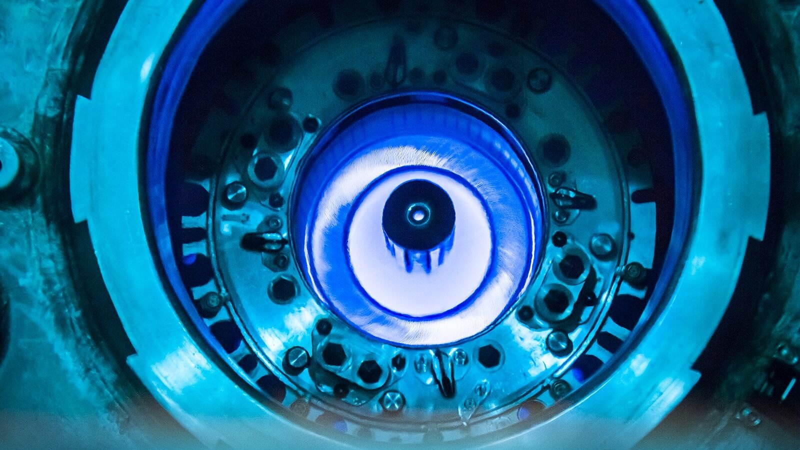 Looking down into a nuclear reactor pool, where the submerged core assembly glows an intense blue against dark metal rings.