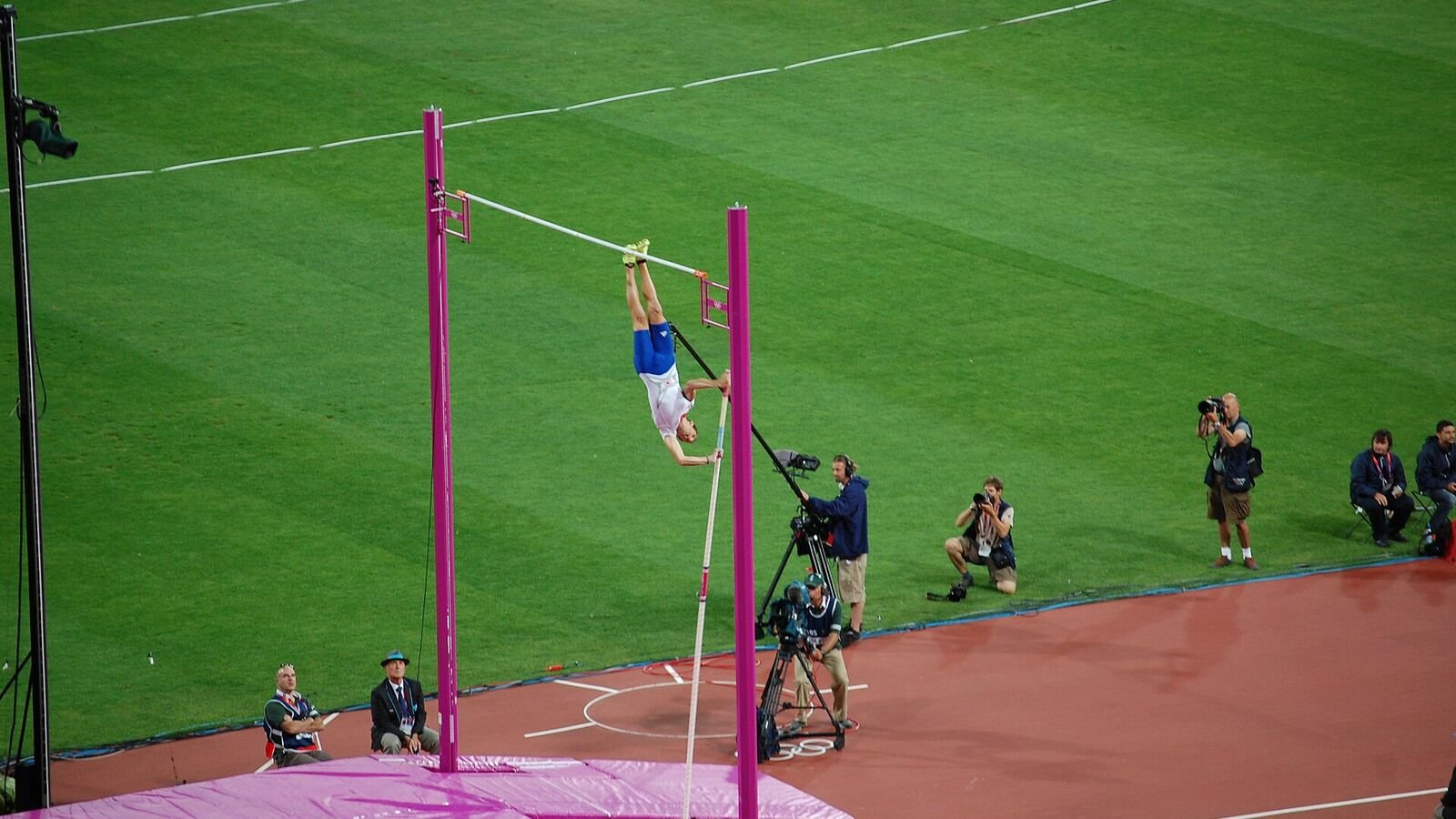 A pole vaulter photographed upside down at the top of a vault, body arched over a raised crossbar with the bent fibreglass pole falling away below.