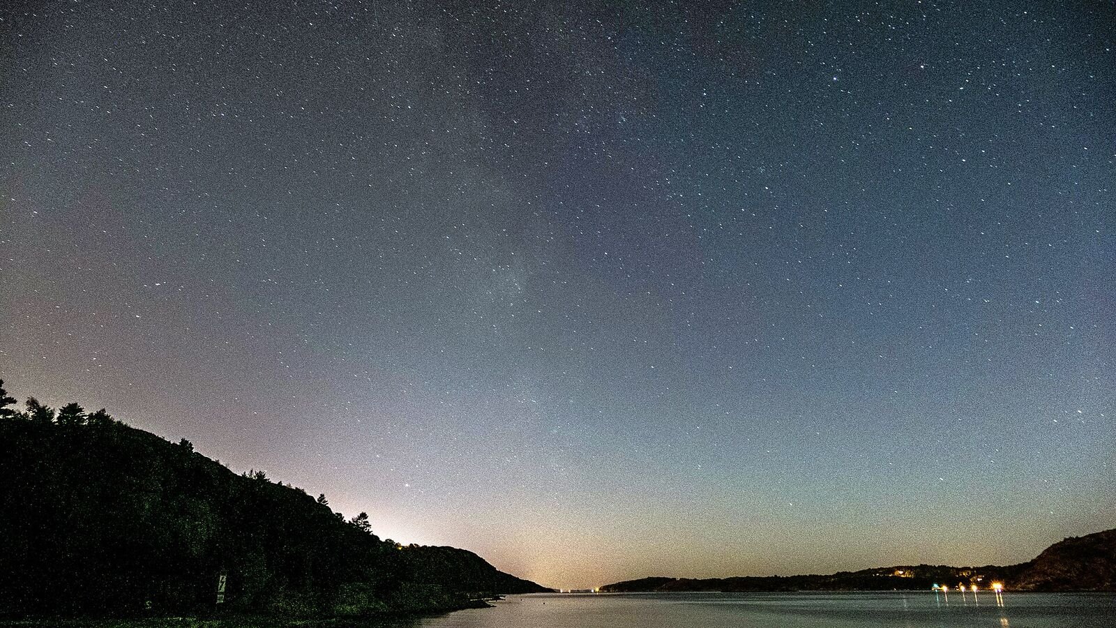The Milky Way arching over a dark fjord, its stars reflected in still water, with a faint orange glow of town lights on the horizon