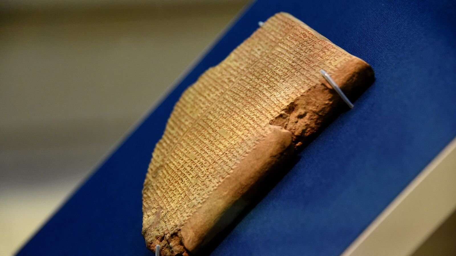 A broken clay tablet covered in rows of small cuneiform script, mounted at an angle on a blue backing board inside a lit museum display case.