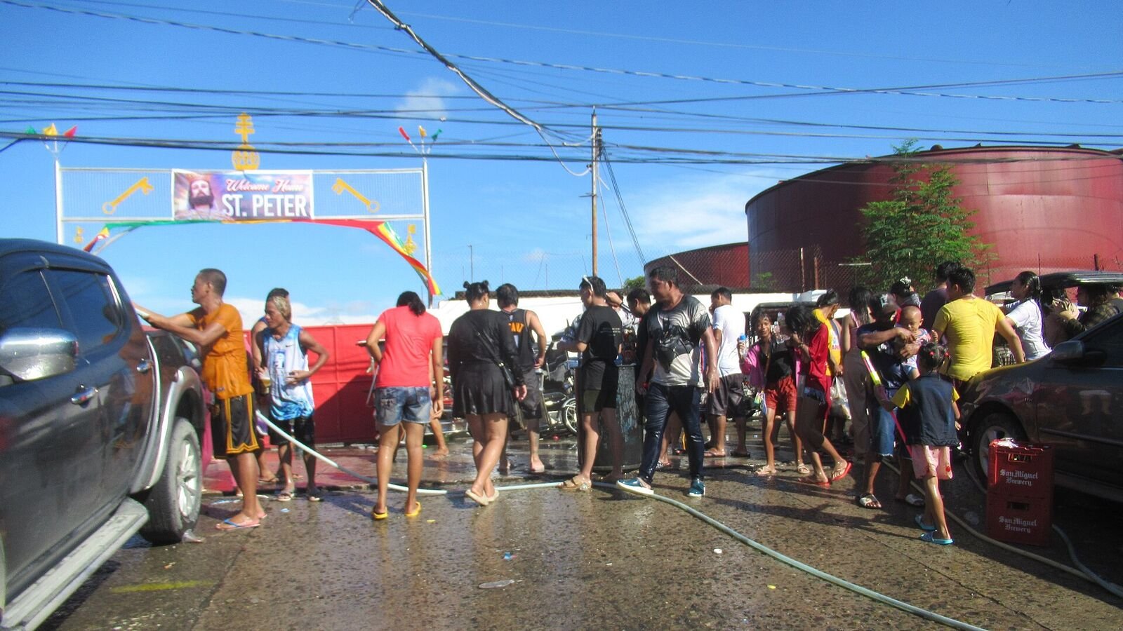 A crowded street under a bright blue sky during a Philippine fiesta, with people spraying water from hoses over one another beside parked vehicles and a decorated arch reading Saint Peter.
