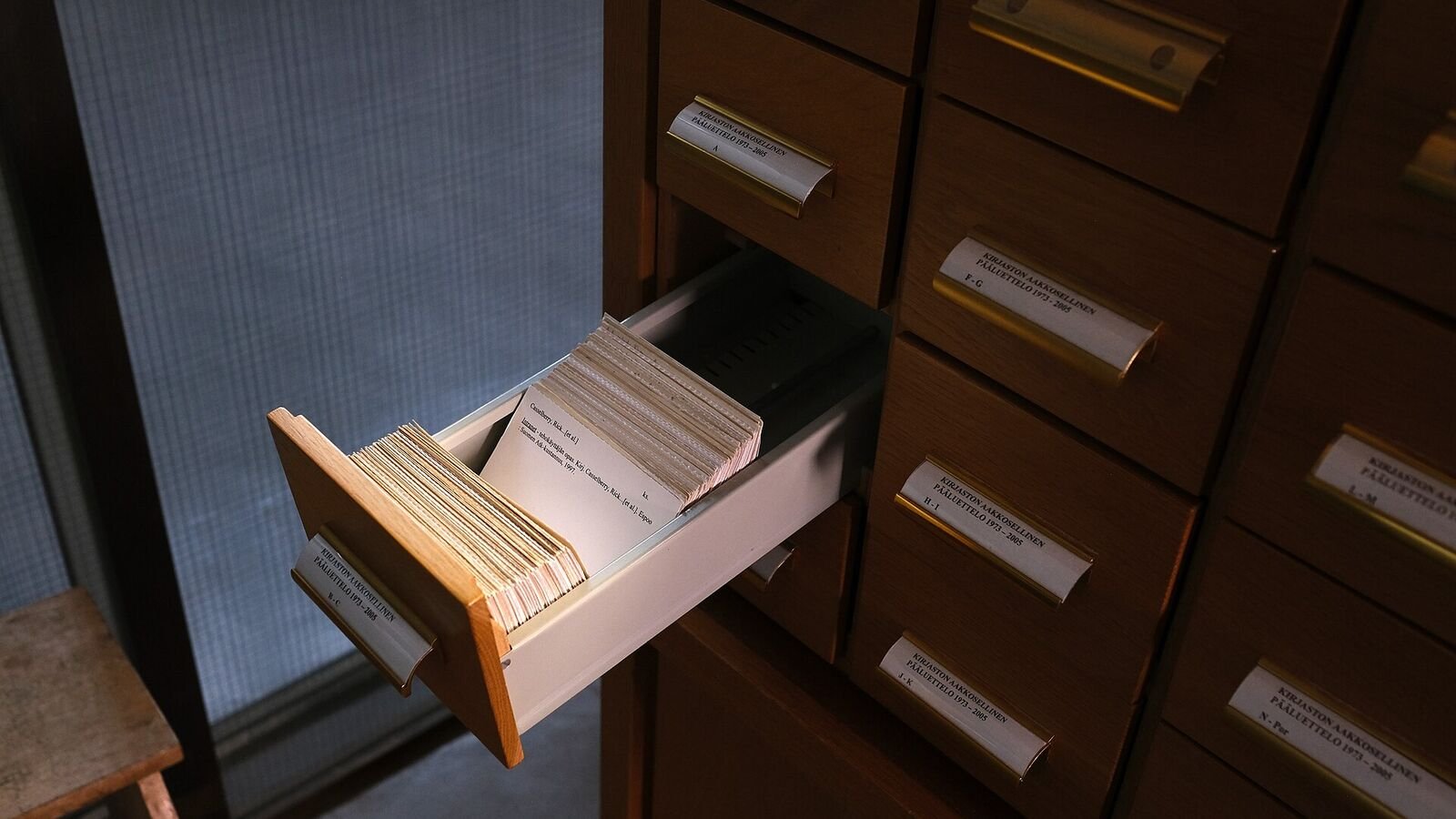 An open drawer in a wooden library card-index cabinet, packed tight with handwritten and typed index cards, with labelled brass handles on the surrounding drawers.