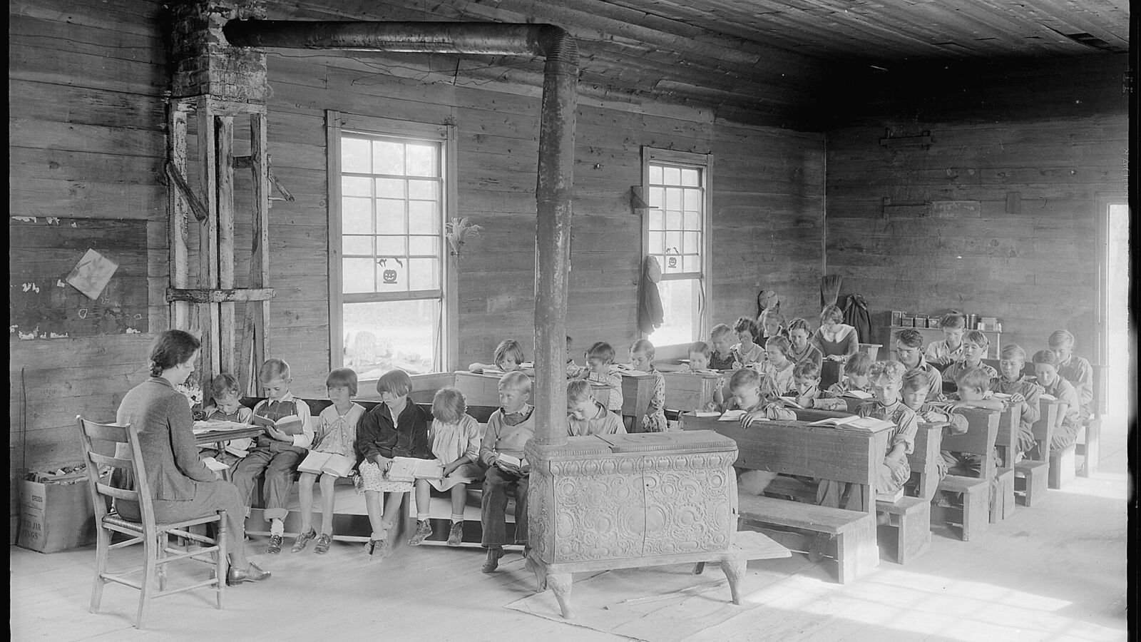 Interior of a rural American schoolroom in the 1930s, with pupils of several ages sitting at wooden desks in rows facing a blackboard.